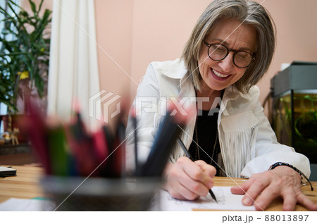 View through blurred color pencil holder at happy smiling senior woman fashion designer creating new clothing line and clothing collection inspired sketching in her creative fashion design studio View through blurred color pencil holder at happy smiling senior woman fashion designer creating new clothing line and clothing collection inspired sketching in her creative fashion design studio 88013897