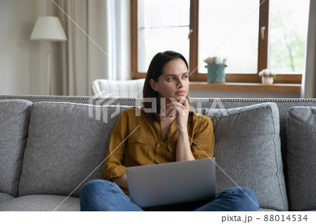 Pensive woman sits on sofa with laptop looking into distance Pensive woman sits on sofa with laptop looking into distance 88014534