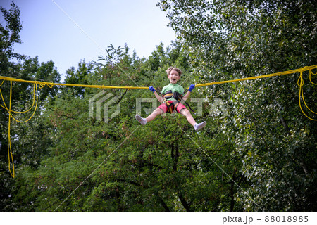 The boy is jumping on a bungee trampoline. A child with insurance and stretchable rubber bands hangs against the sky. The concept of happy childhood and games in the amusement park 88018985