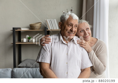Elderly loving couple standing in living room looking out window 88020199