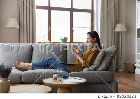 Smiling young woman holds smartphone relaxing on sofa with laptop 88020233