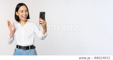 Image of stylish modern asian woman waving hand at smartphone front camera, video chat, chatting with person on mobile phone application, white background Image of stylish modern asian woman waving hand at smartphone front camera, video chat, chatting with person on mobile phone application, white background 88024652