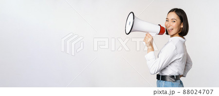 Beautiful young asian woman talking in megaphone, screams in speakerphone and smiling, making announcement, shout out information, standing over white background 88024707