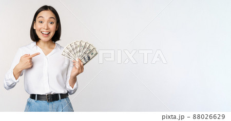 Portrait of young asian woman pointing at her money dollars, showing cash, standing over white background Portrait of young asian woman pointing at her money dollars, showing cash, standing over white background 88026629