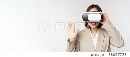 Meeting in vr chat. Asian businesswoman in virtual reality glasses, raising hand and saying hello, greeting someone, standing over white background 88027723