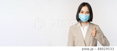 Coronavirus and workplace concept. Portrait of businesswoman in medical face mask showing thumbs up, wearing business suit, standing over white background 88029533
