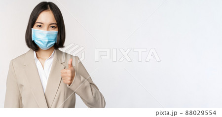Coronavirus and workplace concept. Portrait of businesswoman in medical face mask showing thumbs up, wearing business suit, standing over white background Coronavirus and workplace concept. Portrait of businesswoman in medical face mask showing thumbs up, wearing business suit, standing over white background 88029554