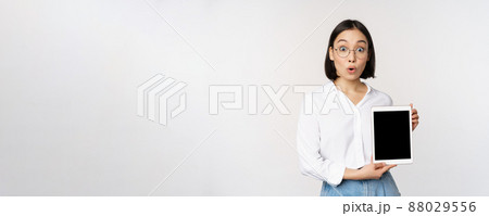 Enthusiastic asian woman, office worker in glasses showing digital tablet screen, demonstrating info on gadget display, standing over white background 88029556