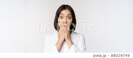Close up face of asian woman gasping, looking shocked and speechless, holding hands near mouth and staring at camera impressed, white background 88029749