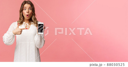 Have you seen it, awesome app. Impressed and excited pretty blond girl in white dress recommend download application, visit online store, pointing at mobile phone and stare camera thrilled Have you seen it, awesome app. Impressed and excited pretty blond girl in white dress recommend download application, visit online store, pointing at mobile phone and stare camera thrilled 88030525