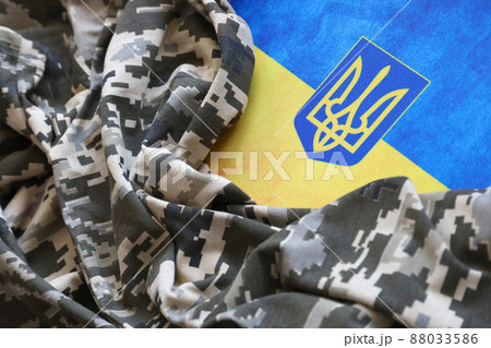 Ukrainian flag and coat of arms with fabric with texture of pixeled camouflage. Cloth with camo pattern in grey, brown and green pixel shapes with Ukrainian trident sign 88033586