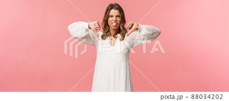 Portrait of picky and moody cute blond girl complaining on bad service, awful quality, show thumbs-down and tongue with dislike grimacing, telling about disgusting experience, pink background 88034202