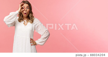 Portrait of amazed and intrigued young blond girl seeing something awesome, look from okay sign with startled excited expression, fold lips say wow, pop eyes at camera, pink background 88034204