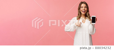 Portrait of upbeat good-looking blond woman in white dress, recommend download app or subscribe, using online shopping application, make thumb-up, show mobile phone screen 88034232