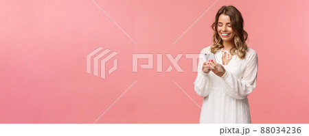 Technology and smartphones concept. Attractive young woman in cute white dress messaging, arrange romantic date, booking tickets via online application on mobile phone, smiling at display 88034236