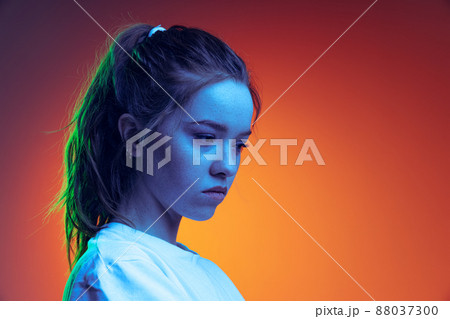 Portrait of young, sad looking girl posing isolated over gradient red orange background in blue neon light Portrait of young, sad looking girl posing isolated over gradient red orange background in blue neon light 88037300