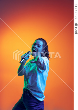 Portrait of young girl emotively singing in microphone, posing isolated over gradient red background in neon light. Music lifestyle Portrait of young girl emotively singing in microphone, posing isolated over gradient red background in neon light. Music lifestyle 88037310