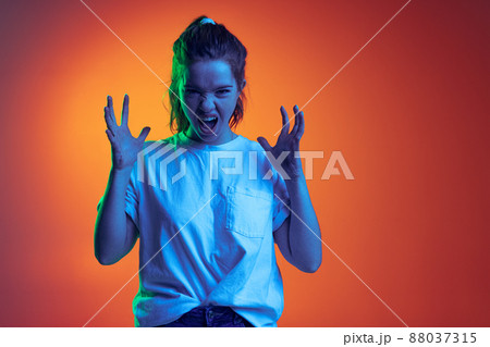 Portrait of young expressive girl in white T-shirt making faces isolated over orange background in neon light Portrait of young expressive girl in white T-shirt making faces isolated over orange background in neon light 88037315