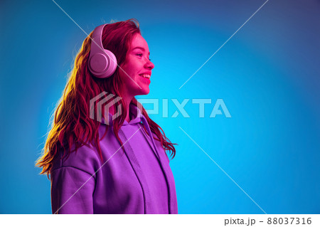 Side view portrait of young girl in white headphones posing isolated over blue studio background in pink neon light 88037316