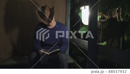 Cowboy man is reading a paper book sitting on the porch of a village house. The concept of reading paper books, learning and hobbies. Dark night. Old, retro decor, burlap background Cowboy man is reading a paper book sitting on the porch of a village house. The concept of reading paper books, learning and hobbies. Dark night. Old, retro decor, burlap background 88037932