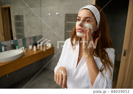 Attractive pregnant red haired woman in white bathrobe standing in the bathroom and taking care of her face skin, applying moisturizer or facial exfoliating scrub, doing smoothing massage movements Attractive pregnant red haired woman in white bathrobe standing in the bathroom and taking care of her face skin, applying moisturizer or facial exfoliating scrub, doing smoothing massage movements 88039052