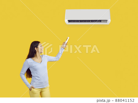 Woman with remote control adjusts comfortable temperature on air conditioner on yellow background. Young happy woman uses modern air conditioning for her comfort. Banner. Copy space. Advertising. 88041152