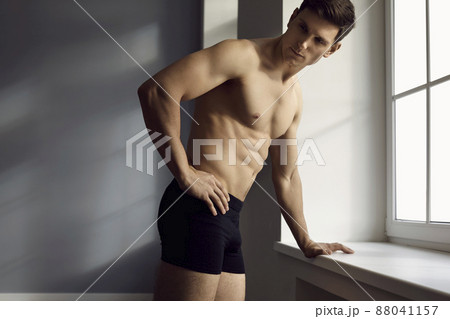 Handsome fit bare chested young man in underwear standing by the window. Male model with strong face and athletic body wearing comfortable black cotton underpants posing in the studio. Fashion concept Handsome fit bare chested young man in underwear standing by the window. Male model with strong face and athletic body wearing comfortable black cotton underpants posing in the studio. Fashion concept 88041157