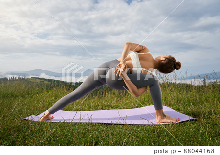 Young woman in sport bra and leggings stretching body on yoga mat with view on mountains. Fitness female person enjoying outdoors activity during summer day. 88044168