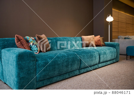 Exhibition of modern stylish upholstered furniture in the showroom of a furniture store. Focus on a turquoise soft velour sofa and brown pillows lit by a lamp against a brown wall background 88046171
