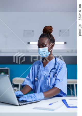 Clinic medical nurse looking for patient disease records and clinical documents while wearing virus protection mask. Hospital staff worker using laptop to search treatment medicine and patient 88048100