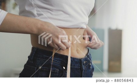 A young woman stands in front of a mirror and measures the circumference of her waist with a tape measure. Close-up 88056843