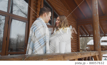 A guy and a girl wrapped in a blanket, standing at a wooden house in winter. Romance 88057632