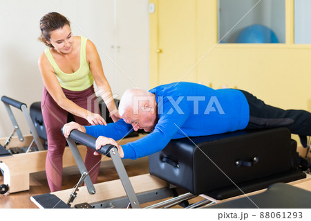 Instructor of pilates helping elderly man workout. Two people working in pilates studio, woman assistant supporting and correcting old man patient beginner. 88061293