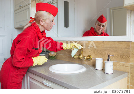 A woman in a red uniform wipes the faucet in the bathroom with a rag. Disinfection in yellow rubber gloves . Cleaning concept. A woman in a red uniform wipes the faucet in the bathroom with a rag. Disinfection in yellow rubber gloves . Cleaning concept. 88075791