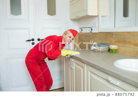A woman in a red uniform cleans the countertop in the bathroom, wiping it with a rag. Disinfection in yellow rubber gloves. A woman in a red uniform cleans the countertop in the bathroom, wiping it with a rag. Disinfection in yellow rubber gloves. 88075792