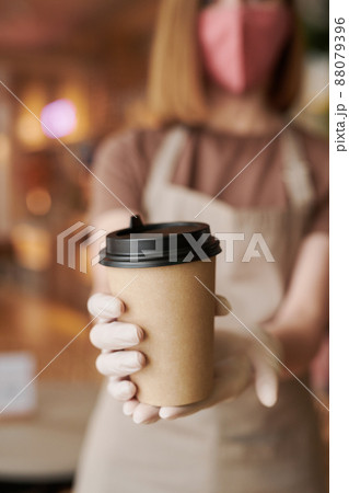 Close-up selective focus on foreground shot of unrecognizable waitperson wearing mask and gloves giving cup of coffee to customer Close-up selective focus on foreground shot of unrecognizable waitperson wearing mask and gloves giving cup of coffee to customer 88079396
