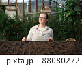 Young woman stands near table with seedlings in large greenhouse and holds rake and shovel in her hands. Gardening, seedlings, sales 88080272