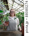 Young woman stands near table with flowers in the greenhouse and holds pot with green flower. Gardening, seedlings, sale 88080274
