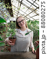 Young woman stands near table with flowers in the greenhouse and holds pot with green flower. Gardening, seedlings, sale 88080275