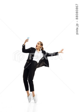 Dynamic portrait of young woman, office worker in black official suit having video call on phone isolated over white studio background 88080867
