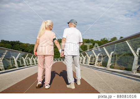 Positive senior woman and man walk holding hands along footbridge after training Positive senior woman and man walk holding hands along footbridge after training 88081016