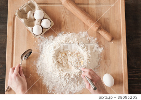 Above view of unrecognizable woman mixing flour and egg with whisk on wooden board with rolling pin Above view of unrecognizable woman mixing flour and egg with whisk on wooden board with rolling pin 88095205