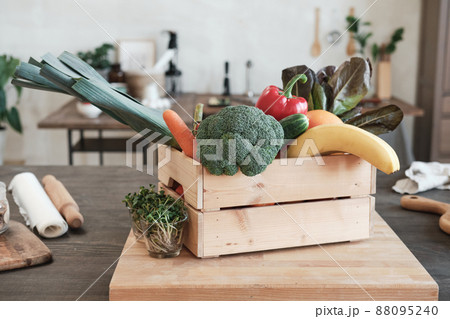 Wooden box full of various vegetables and fruits placed on board with growing plants into glasses in modern kitchen 88095240
