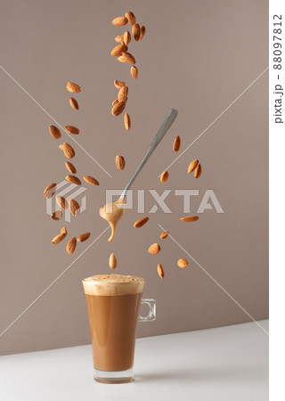Glass cup with cappuccino and fragrant foam and a spoon and falling almonds. Cup with cappuccino, latte or coffee with milk. Creative design for cafes and restaurants with a cup of coffee.  88097812