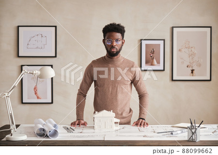 Portrait of serious confident African American architect in round-shaped eyeglasses standing at table with blueprints in office 88099662