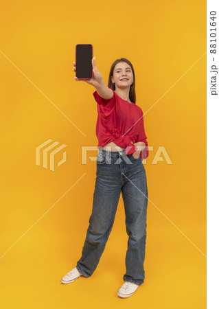 Teenage girl in red blouse holds phone with blank screen. Full-length portrait of girl with smartphone on yellow background. Copy space. Mock up. Teenage girl in red blouse holds phone with blank screen. Full-length portrait of girl with smartphone on yellow background. Copy space. Mock up. 88101640