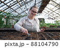 Young woman stands near table with seedlings in large greenhouse and holds rake and shovel in her hands. Gardening, seedlings, sales 88104790