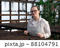 Young woman stands near table with seedlings in large greenhouse and holds rake and shovel in her hands. Gardening, seedlings, sales 88104791