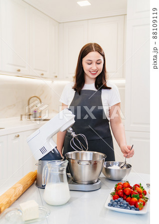 A young woman cook adds a spoonful of flour to prepare dough in a mixer bowl. A young woman cook adds a spoonful of flour to prepare dough in a mixer bowl. 88109119