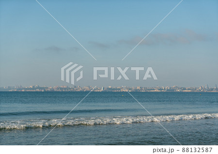 Sea waves running on a sandy shore on a clear sunny day. City in the background. Sea waves running on a sandy shore on a clear sunny day. City in the background. 88132587